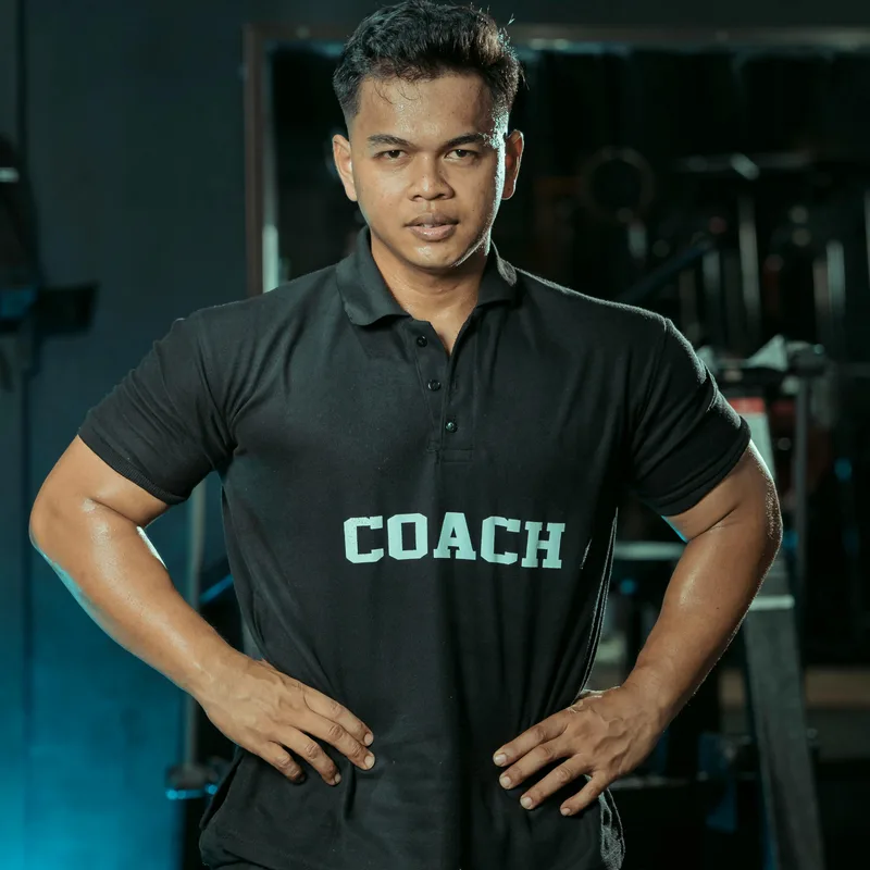 A fitness coach in a black polo shirt printed with the word COACH, hands on hips in a darkened gym