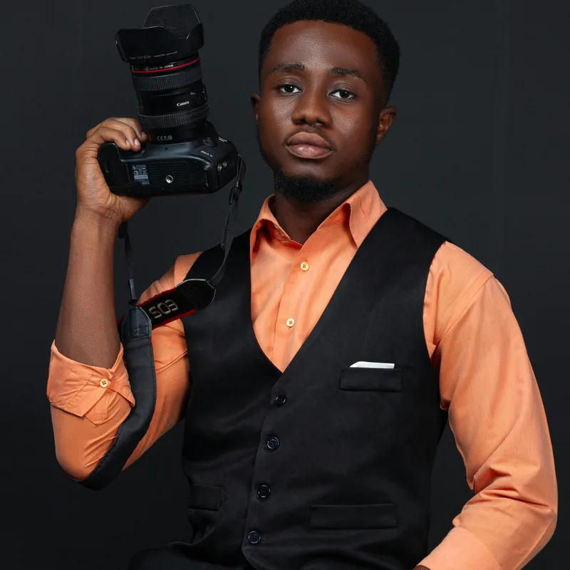 A freelance photographer in an orange shirt and black waistcoat holding a Canon DSLR camera beside his head, against a dark studio backdrop