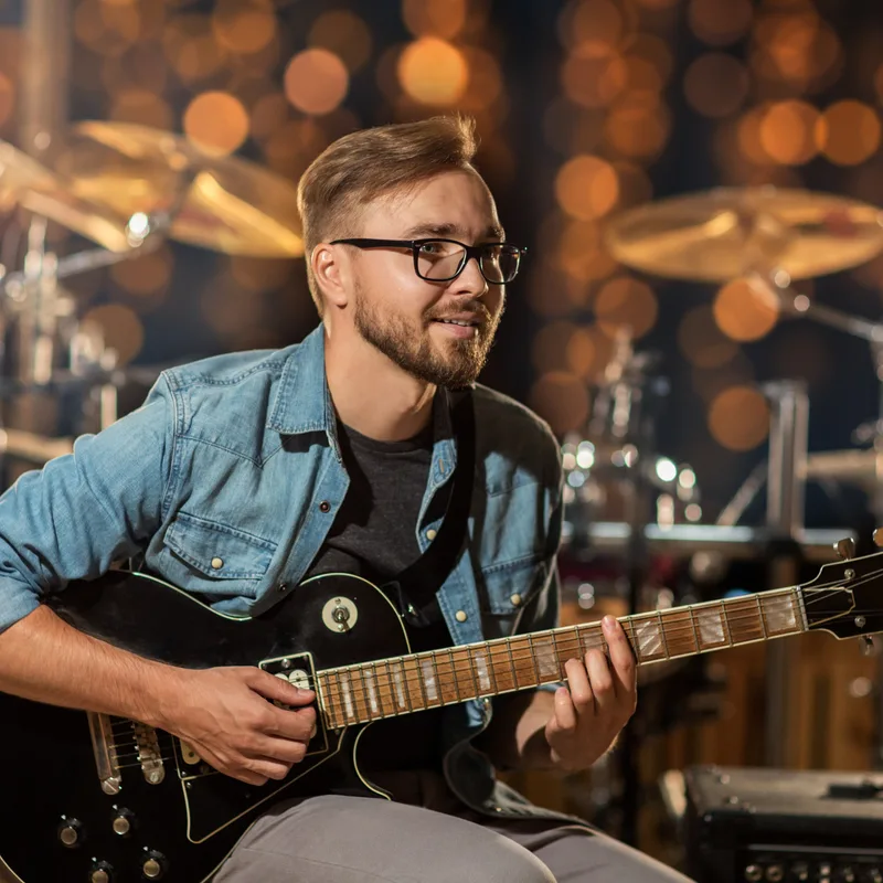 A musician in a denim shirt playing a black electric guitar on a dim stage, with a drum kit and warm bokeh lights behind him